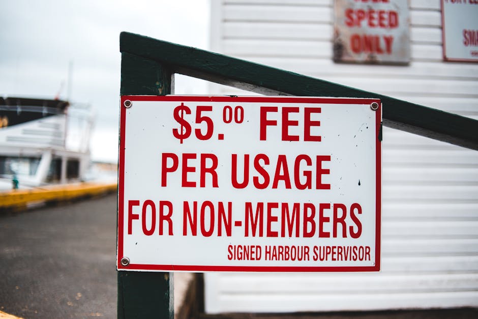Close-up image of a rectangular white sign with bold red text displaying a fee of $5.00 per usage for non-members, attached to a dark green railing at a harbour or marina. In the background, blurred boats and a paved dock area are visible, suggesting a waterfront environment. The sign, signed by the harbour supervisor, indicates a charge applicable for accessing or using certain facilities or services, possibly related to boat storage or maintenance. The scene appears outdoors, under overcast weather, with the sign prominently positioned to inform visitors or clients about the cost associated with harbour access. This setting relates to logistical aspects of house and vessel removals or transportation, highlighting the importance of clear fee signage in commercial or service environments involved in relocations or transport logistics, similar to professional removals by companies like Man and Van Knightsbridge.