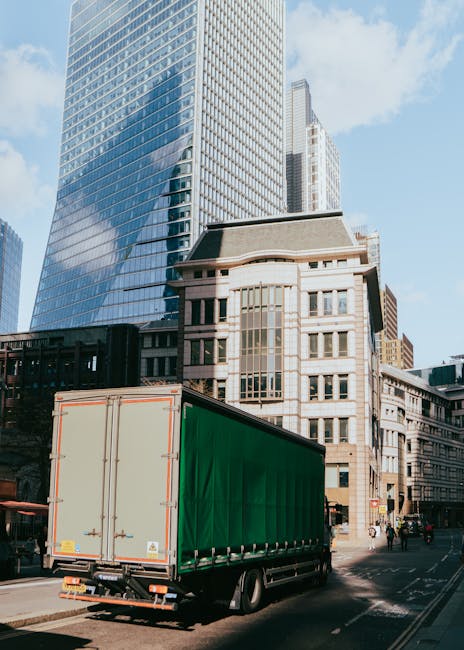 A large green commercial delivery truck parked on a city street surrounded by tall, modern office buildings with glass facades and older stone structures. The truck is positioned near the curb, ready for loading or unloading, with its rear doors closed. The street appears busy with some pedestrians and vehicles visible in the background. The scene is captured during daylight with clear skies. The setting reflects urban logistics typical of house removals and furniture transport services, with the truck potentially being used by Man and Van Knightsbridge for home relocation and moving processes in a busy city environment.