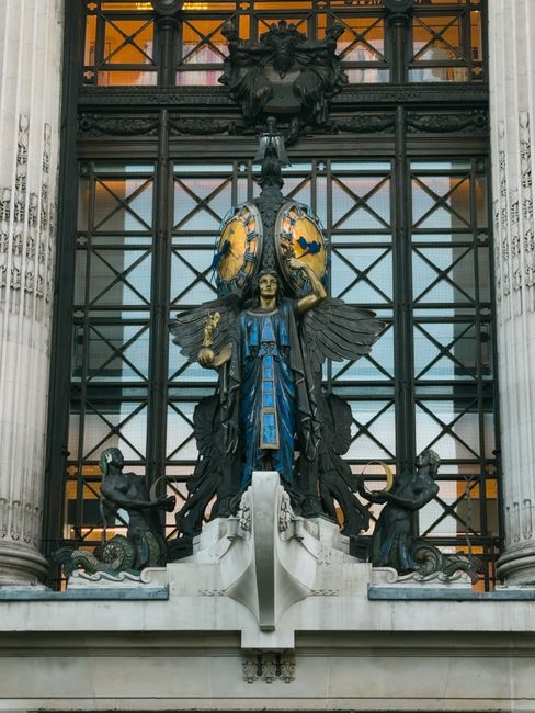 The image shows the ornate façade of a historic building with intricate architectural details. Prominently centered is a large bronze sculpture featuring a winged female figure dressed in a blue gown, holding a circular shield with yellow and blue designs. The sculpture is positioned above the entrance, which is framed by tall, classical columns. Above the sculpture, there is a decorative iron grille with geometric patterns and an elaborate crest or emblem at the top. The lighting appears natural, illuminating the craftsmanship of the sculpture and architectural elements. This scene is suggestive of London's historic cityscape, fitting for a context related to house removals, furniture transport, or moving logistics, such as those provided by Man and Van Knightsbridge. The building's detailed façade and the decorative historic elements highlight the importance of careful handling during home relocation or professional removals, aligning with services in the Knightsbridge area.