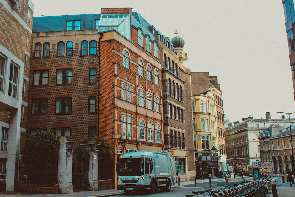 A street scene in Knightsbridge, featuring several multi-storey buildings with a mix of brick and ornate architectural details, some with arched windows and decorative elements, indicating a historic area. In the foreground, a white man with a high visibility vest is loading large cardboard boxes and wrapped furniture onto a light blue van parked on the pavement, which is part of a home relocation or furniture transport operation by Man and Van Knightsbridge. The loading process involves the use of a trolley to move boxes and larger items from the building into the vehicle, with additional packing materials such as plastic wrap visible. Surrounding the van, other moving equipment, including straps and blankets, are arranged to facilitate safe handling of items. The scene takes place during daylight with natural lighting, and the street is lined with bollards and parked bicycles, with nearby buildings and street lamps completing the urban environment, reflecting an organized and professional removals process in accordance with local house removals procedures.