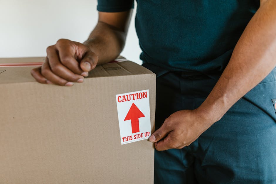 A person wearing a dark green shirt and navy trousers is holding a cardboard moving box, which features a red and white caution label indicating 'This Side Up' with an upward pointing arrow. The individual is gripping the box with one hand on the top edge and the other on the side, demonstrating proper handling during a home relocation process. The scene appears to be inside a property, possibly near an entrance or corridor, with the background showing neutral-colored walls. The cardboard box is sealed with packing tape and is likely being prepared for loading onto a van or for transport within the house as part of packing and moving activities. The setting emphasizes careful handling of items, an essential aspect of professional removals by companies such as Man and Van Knightsbridge, especially during furniture transport and loading procedures for house removals.