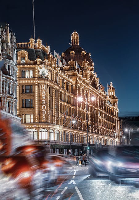 Nighttime exterior view of the Harrods department store in Knightsbridge, illuminated with numerous bright decorative lights outlining its ornate architectural features, including domes and turrets. The building's signage clearly displays 'HARRODS' in large lit letters on a vertical sign. In the foreground, a busy city street shows blurred motion of passing cars, indicating movement and traffic, with some pedestrians walking along the pavement near the building. The scene captures the lively atmosphere of central London, with the illuminated facade providing a striking contrast against the dark evening sky. This image reflects the urban environment associated with house removals and furniture transport services, emphasizing the importance of logistical planning during home relocations, especially in a prominent area like Knightsbridge. Man and Van Knightsbridge services support smooth moves through careful packing and transportation of household items within this bustling cityscape.