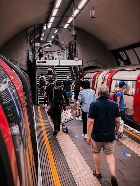 Image of passengers disembarking from a red and white London Underground train inside an curved underground station tunnel with a tiled ceiling and modern lighting. Several individuals are walking along the platform, some carrying luggage or backpacks, while others are descending a staircase towards the station's upper level. The platform surface features tactile paving strips and yellow safety lines, indicating a busy transit environment. The scene depicts a typical morning or daytime commute, with people transitioning between train and station access points. This setting exemplifies the logistical environment encountered during property or furniture transport, involving careful movement and handling of belongings in an urban public transport context, relevant to home relocation or removals.