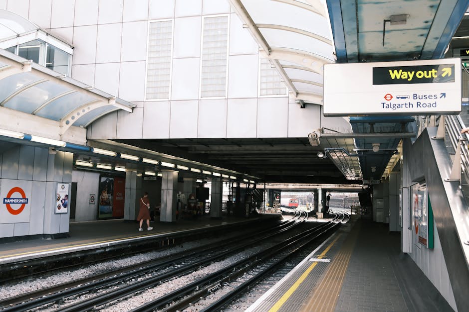 Exterior view of Knightsbridge station entrance in daytime, featuring the railway tracks and platform area with a curved shear wall and overhead canopy. The station walls are covered with metallic panels, and a yellow 'Way out' sign with an arrow points toward the exits. To the left, a person dressed in casual clothing is walking along the platform, while a small group of people is visible in the background near the entrance/exit area. The platform surface includes tactile paving for safety, and the station provides access to bus services on Talgarth Road. This setting illustrates the typical environment for home relocation and furniture transport, with a focus on passenger and baggage movement, relevant to moving and removals services like those offered by Man and Van Knightsbridge.