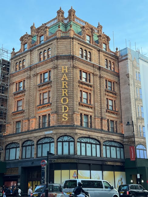 A large multi-storey building with ornate architectural details, situated on a busy urban street. The facade features numerous rectangular windows with decorative frames, and the top of the building is crowned with domed and spire-like features. The word 'HARRORDS' is displayed vertically in large gold letters on the upper part of the building. The ground floor has large arched windows, possibly storefronts, with dark framing. In front, vehicles including a white van, cars, and a bicycle are visible, along with pedestrians on the pavement. A man and a woman appear to be loading or unloading items near the van, with cardboard boxes, plastic wrapping, and packing materials nearby. The scene is well-lit with natural daylight, and neighboring buildings with similar architectural styles are partially visible. This setting suggests a building undergoing or associated with home relocation or furniture transport, aligned with professional removals and moving services.