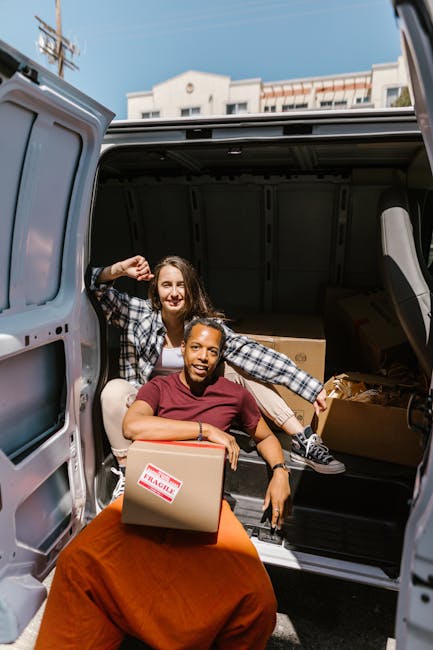 Two women are engaged in a home relocation process, loading as well as unloading cardboard boxes filled with household items into the back of a large, grey moving van. The van's rear doors are wide open, revealing a spacious interior stocked with additional boxes and protective packaging materials such as plastic wrap and blankets, which are used to safeguard furniture during transport. One woman, seated at the edge of the van's open hatch, is holding a medium-sized box with a 'FRAGILE' sticker, while the other woman, standing inside the vehicle, is lifting and placing another box onto a stack inside. The scene takes place outdoors on a paved street with buildings and satellite dishes visible in the background under a clear sky, with natural daylight illuminating the area. The moving process is in progress, highlighting the careful handling of household belongings typical of professional removals services offered by Man and Van Knightsbridge, supporting efficient furniture transport and packing during a house move.