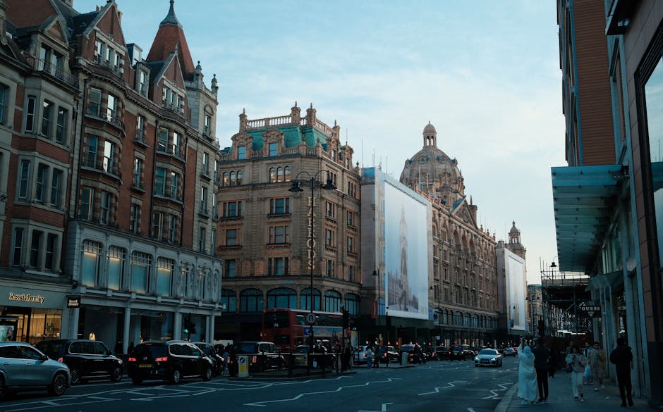 A large multi-storey building with ornate architectural details, situated on a busy urban street. The facade features numerous rectangular windows with decorative frames, and the top of the building is crowned with domed and spire-like features. The word 'HARRORDS' is displayed vertically in large gold letters on the upper part of the building. The ground floor has large arched windows, possibly storefronts, with dark framing. In front, vehicles including a white van, cars, and a bicycle are visible, along with pedestrians on the pavement. A man and a woman appear to be loading or unloading items near the van, with cardboard boxes, plastic wrapping, and packing materials nearby. The scene is well-lit with natural daylight, and neighboring buildings with similar architectural styles are partially visible. This setting suggests a building undergoing or associated with home relocation or furniture transport, aligned with professional removals and moving services.