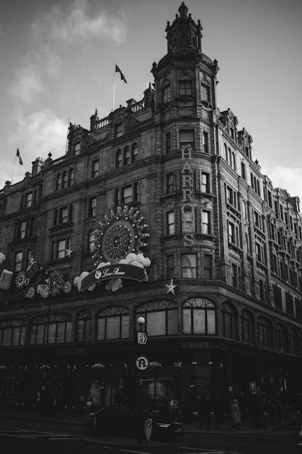 A black-and-white photograph depicts an ornate multi-storey building on a city street, featuring decorative architectural details such as arched windows, intricate stonework, and a prominent corner tower with a flag flying atop. The ground floor houses a venue or establishment with illuminated signage and decorative elements, including a large circular light display and a marquee. In the foreground, a loading area or pavement is visible with a vehicle parked nearby, suggesting a home relocation or furniture transport process. While no moving activity is directly shown, the scene's urban setting and architecture are consistent with a typical city environment where house removals and home relocations could occur. The overall atmosphere emphasizes the historic and architectural character of the building, with some construction or loading equipment potentially nearby, supporting the context of professional removals and packing and moving services carried out by companies such as Man and Van Knightsbridge.
