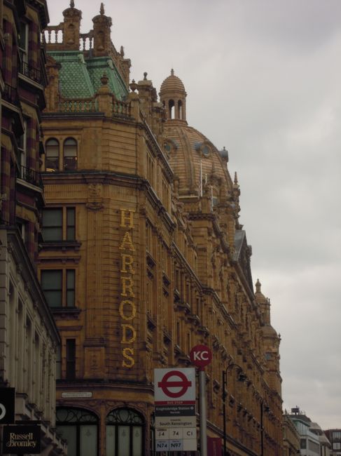 A close-up view of a historic Victorian-style building in Knightsbridge, featuring intricate stonework, decorative columns, and a prominent sign reading 'HARRIERDS' on its façade. In the foreground, a London Underground station entrance with a red circular sign displaying the roundel symbol and a white sign beneath showing bus routes and station information is visible. The sky above is overcast with grey clouds, suggesting a typical London day. This image captures the architectural character of central London and the urban environment where house removals and furniture transport services by Man and Van Knightsbridge may operate, illustrating the type of urban setting involved in home relocation efforts.