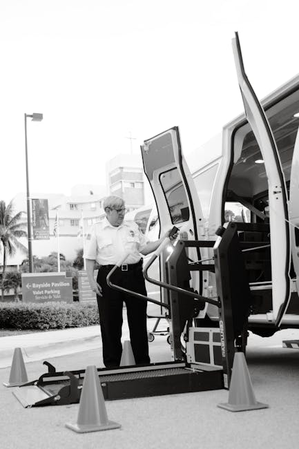 A man wearing glasses and a light-colored shirt is standing beside a white van with its side door open, situated on a paved street. He is holding the handle of a hydraulic lift that extends from the van, which is being used to load or unload large, framed items or furniture wrapped in protective material. The scene includes orange traffic cones arranged around the lifting area to ensure safety. In the background, there are buildings, palm trees, and a streetlamp, indicating an urban setting. The interior of the van shows a glimpse of storage space with packing materials and equipment, consistent with professional removals or moving services. This setup reflects a home relocation or furniture transport process handled by Man and Van Knightsbridge, emphasizing careful packing, loading, and transport logistics for house removals in the area.