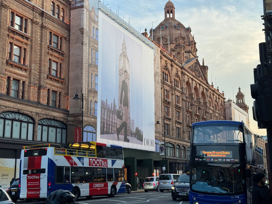 A close-up view of a historic Victorian-style building in Knightsbridge, featuring intricate stonework, decorative columns, and a prominent sign reading 'HARRIERDS' on its façade. In the foreground, a London Underground station entrance with a red circular sign displaying the roundel symbol and a white sign beneath showing bus routes and station information is visible. The sky above is overcast with grey clouds, suggesting a typical London day. This image captures the architectural character of central London and the urban environment where house removals and furniture transport services by Man and Van Knightsbridge may operate, illustrating the type of urban setting involved in home relocation efforts.